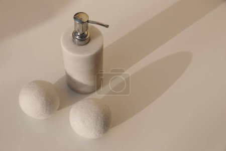 Dryer wool balls and white luxury marble stone soap dispenser bottle on blurred beige table background in sunlight. Long soft shadows. Hygiene, bodycare laundry or bathroom concept, high angle view