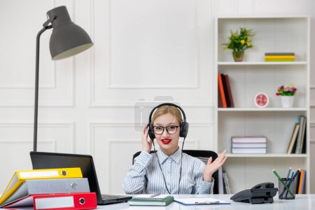 Foto de Secretaria de trabajo en la oficina encantadora linda chica rubia en camisa con carga de trabajo en los auriculares - Imagen libre de derechos