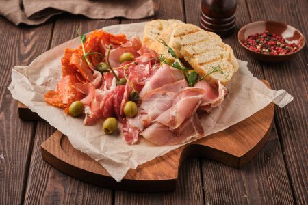 Photo for A close-up view of a gourmet meat platter, featuring sliced ham and salami, garnished with olives and rosemary, accompanied by sliced bread and assorted spices. The meats are artfully arranged on a wooden board placed on a rustic wooden table. A pepp - Royalty Free Image