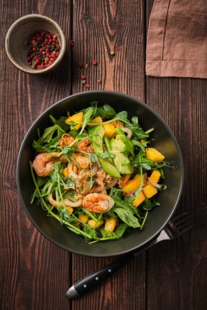 A vibrant seafood salad mixed with fresh greens, juicy mango pieces, and creamy avocado slices, served in a black bowl on a rustic wooden table. The image conveys a mood of freshness and wholesome nutrition.