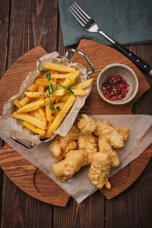 A close-up view of a delicious serving of crispy fish and golden fries. The meal is presented on a wooden board with a small bowl of mixed spices. A fork rests on a dark green napkin beside the meal, all placed on a rustic wooden table.