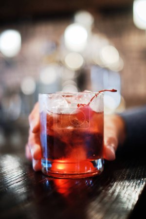 A close-up image capturing a moment of someone holding a glass of cherry cocktail, garnished with a cherry, at a well-lit bar.