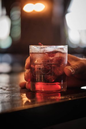 A close-up image capturing a moment of someone holding a glass of cherry cocktail, garnished with a cherry, at a well-lit bar.