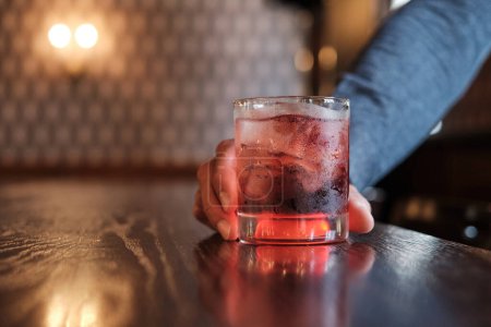 Una imagen de cerca capturando un momento de alguien sosteniendo una copa de cóctel de cereza, adornado con una cereza, en un bar bien iluminado.