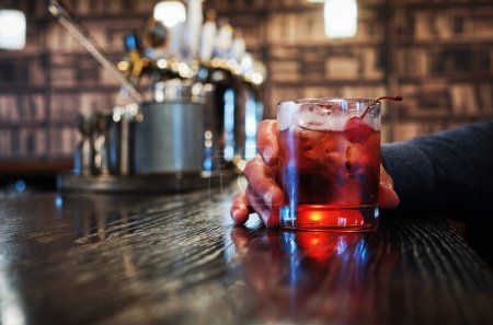 Close-up view of a persons hand holding a glass of red cocktail with ice cubes. The individual is seated at a polished wooden bar, creating a relaxed and cozy atmosphere.