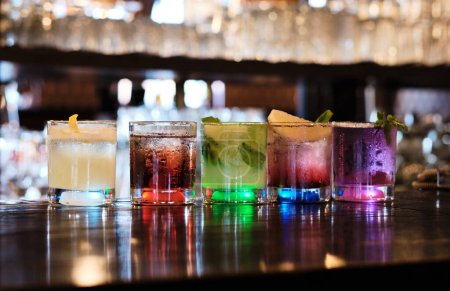A variety of five colorful cocktails, each uniquely garnished, are lined up on a dark wooden bar counter, illuminated from below. The glasses reflect ambient light, creating an elegant and inviting atmosphere.