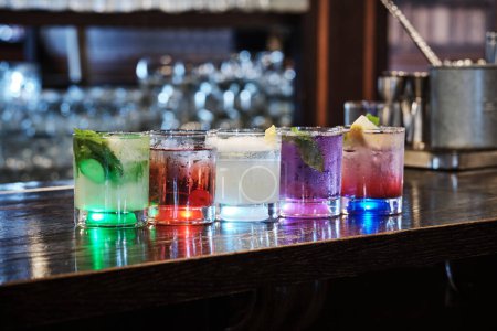 A variety of five colorful cocktails, each uniquely garnished, are lined up on a dark wooden bar counter, illuminated from below. The glasses reflect ambient light, creating an elegant and inviting atmosphere.