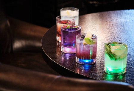 A variety of colorful cocktails with garnishes are displayed on a dark, textured wooden table, illuminated by soft lighting.