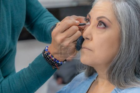 Foto de Stylist putting black eyeliner on lower eyelid in professional makeup session in a senior adult woman, long gray hair, warm colors, lips with pink lipstick, beauty salon. Fashion and beauty concept - Imagen libre de derechos