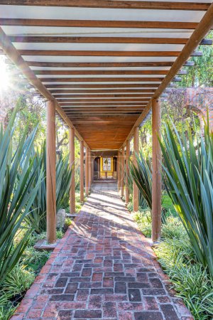 Langer Steinweg im Korridor mit Säulen aus Holzpfählen mit geschlossener Decke inmitten riesiger grüner Pflanzen mit einem Tor im Hintergrund, sonniger Wintertag auf einer Ranch in Mexiko