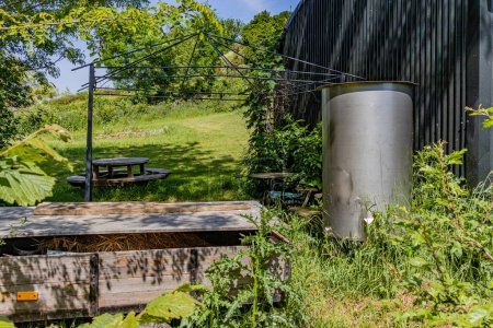 Accumulation of objects in backyard of a farm with wild vegetation on hill in background, camping table, metal tank to store water, metal structure of a tent and wooden box with straw, sunny day