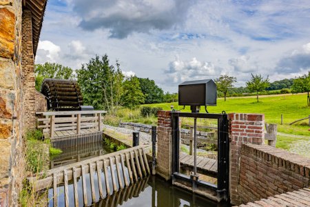 Foto de Puerta cerrada del canal de agua en el antiguo molino de agua Eper o Wingbergermolen, al lado del río Geul, llanura con árboles contra el cielo cubierto de nubes en el fondo, día soleado en Terpoorten, Epen, Limburgo Meridional, Países Bajos - Imagen libre de derechos