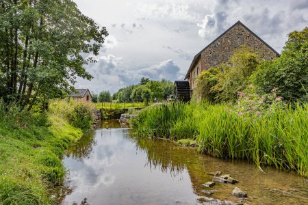 Photo for River Geul with old Eper watermill or Wingbergermolen in background, surrounded by wild vegetation and trees against cloud-covered sky, cloudy day at Terpoorterweg, Epen, South Limburg, Netherlands - Royalty Free Image