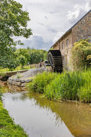 Photo for Vertical shot of Geul river among wild grass with old Eper or Wingbergermolen water mill in background,  trees against cloud covered sky, cloudy day in Terpoorten, Epen, South Limburg, Netherlands - Royalty Free Image