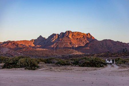 Mexican desert landscape with juniper trees and scrubland, imposing mountain rock formation illuminated by sun in background, car parked on sand, sunset in desert of Baja California Sur, Mexico