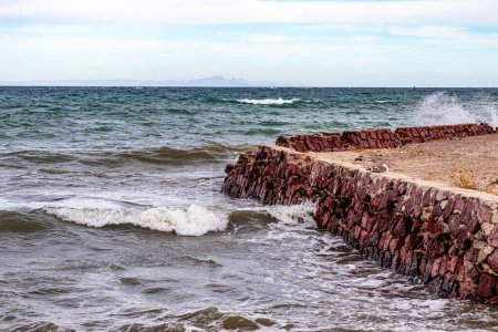 Waves breaking at rustic tourist viewpoint with red stone wall and dirt ground next to seafront, Sea of Cortez and horizon in background, cloudy day in La Paz, Baja California Sur Mexico