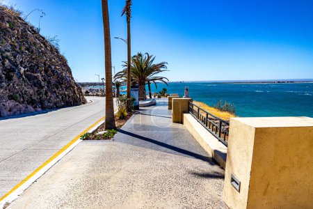 Boardwalk next to Federal Highway 11 and rocky hillside, palm trees at Los Delfines viewpoint against blue sky, sea and horizon in background, sunny day in La Paz, Baja California Sur Mexico