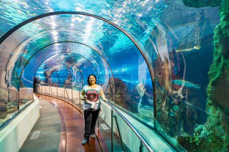 Latin American chubby smiling female tourist standing under glass tunnel of big pond, casual clothes, black hair, blue water in blurred background, Barcelona city aquarium, Spain