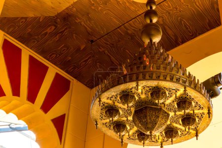 Photo for Arabic or Moroccan style bronze lamp hanging from ceiling, replica of Cordoba mosque building, yellow walls and arched entrance with red decoration in background - Royalty Free Image