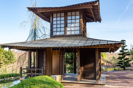 Replica of small wooden Japanese temple next to stream against blue sky, Japanese pavilion in Dutch public park, sunny morning day in Landgraaf, South Limburg in the Netherlands