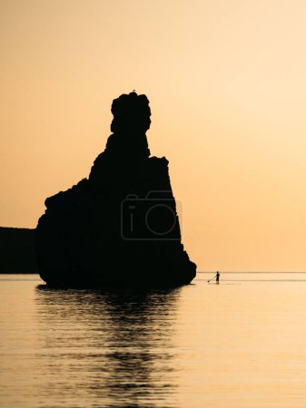 Silhouette of a man floating on a SUP board with an oar and big stone rock. Dawn sun on the sea. Swimming on a supboard at sunset. Active water sports and beautiful landscape of Ibiza