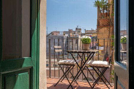 View from the apartment of typical Spanish old house with beautiful balcony with wooden garden furniture, plants and flowers in summer sunny day. View of Barcelona through open wooden shutters