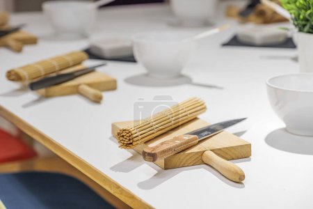 Téléchargez les photos : Ensemble de couteaux, planches et tapis de sushi en bambou sur table blanche avant le cours de maître de sushi. Éducation, cuisine et divertissement concept - en image libre de droit