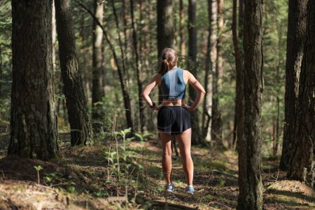 Rear view of female trail runner dressed in sportswear standing on trail in the forest and preparing to run