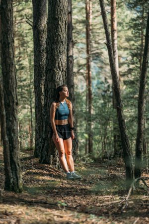 Tired young woman in sportswear leaned on a tree and resting in the forest after working out. Trail running and active life concept