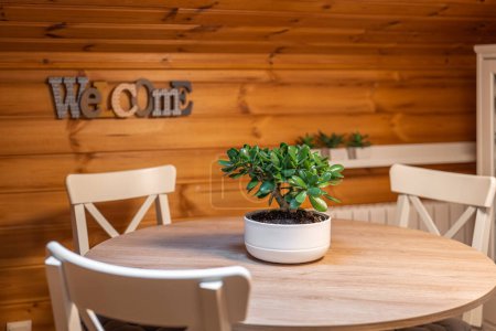 Foto de Planta de jade sobre mesa de comedor redonda con sillas y palabra de bienvenida en el fondo en la pared de madera en una casa rural. Enfoque selectivo - Imagen libre de derechos
