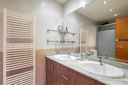 Bathroom with bright light from ceiling lights, beams of cold light reflecting on shiny white surfaces of sinks and in large clean mirror which shows wooden closed door shower cabin heating radiator