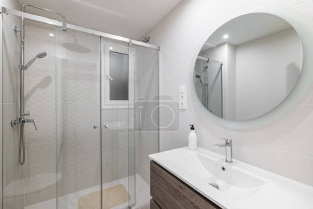 Photo for White ceramic tile bathroom with large round mirror on the wall reflecting the shower area with glass railing and metal fittings - Royalty Free Image