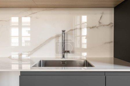 Modern small square sink in the kitchen. Fashionable built-in stainless steel sink. Interior kitchen in white and gray colors with marble tiles on the wall. White countertop and gray kitchen cabinet