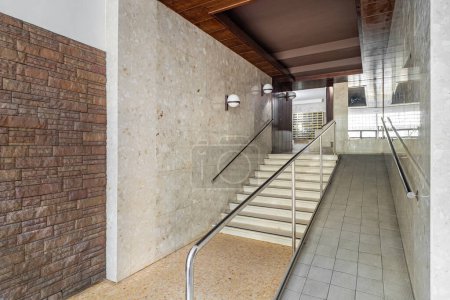 Eingang eines Hauses mit langem Flur, Treppe zum Aufzugsbereich mit Rampe für Rollstuhlfahrer, gemauerte und geflieste Wände, Metallbriefkästen