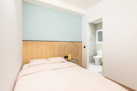 Photo for Double bed against a shallow niche with a blue wall and wooden decor. In the bedroom, there is an open door overlooking a bright toilet room with a toilet bowl and a small plastic window - Royalty Free Image