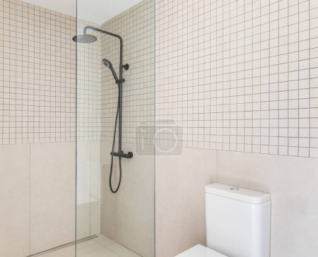 Small bathroom with white toilet with closed lid and an enclosed shower area with transparent glass wall. Walls made of beige tiles of different shapes are harmoniously combined with each other