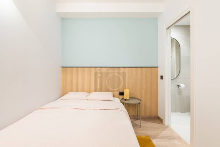 Photo for Double bed against a shallow niche with a blue wall and wooden decor. In the bedroom, there is an open door overlooking a bright toilet room with a toilet bowl and a small plastic window - Royalty Free Image