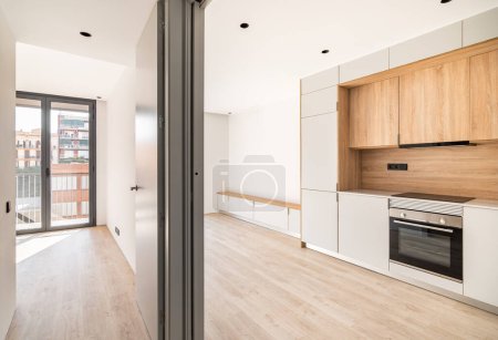 Empty contemporary kitchen, living room and bedroom with balcony at refurbished apartment. Wooden furniture and modern appliances.