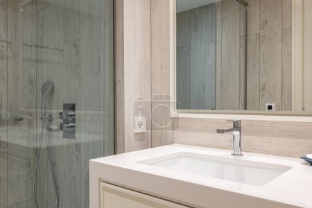 Helle Badezimmer-Nahaufnahme mit Holz-Effekt-Paneelwänden, quadratischem Keramikwaschbecken mit Wasserhahn auf Marmorarbeitsplatte und Spiegel an der Wand. Duschbereich ist von einer gläsernen bläulichen Wand umgeben