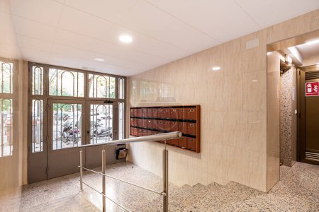 Foto de Vestíbulo en la entrada de la casa con pasillo largo, escaleras al ascensor, baldosas de granito, paredes beige con muchos buzones marrones - Imagen libre de derechos