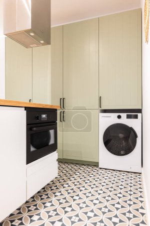 Part of spacious kitchen with modern appliances with black elements. Large built-in wooden cabinet for kitchenware and utensils in beige color with black handles. Floor is made of marble mosaic tiles
