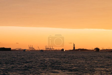 Scenic image capturing the Statue of Liberty with a beautiful sunset backdrop over the New York City harbor. The sky is lit with warm hues, and boats are visible on the water.