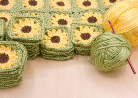 Close up on piles of finished crochet granny squares, connected squares behind with balls of yellow and green yarn to side on light wood table. Crochet hook in green yarn ball.