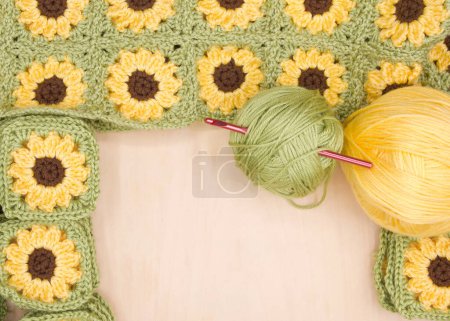 Close up on piles of finished crochet granny squares, connected squares behind with balls of yellow and green yarn to side on light wood table. Crochet hook in green yarn ball. View from above