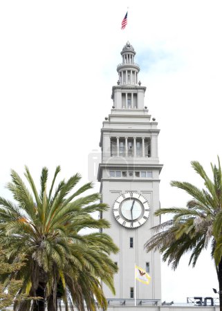 Icónica torre del reloj del Puerto de San Francisco Ferry Building vista a través de palmeras con cielo nublado detrás.