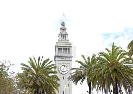 Iconic clock tower of San Francisco Port Ferry Building seen through Palm Trees with cloudy sky behind.