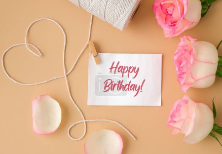 Foto de Tender pink roses with spool of white cotton rope in heart shape on beige background. Paper note with text HAPPY BIRTHDAY. Neutral earth tones. Greeting card holiday. Romantic pastel pink rose flowers - Imagen libre de derechos