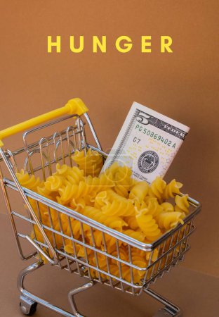 Téléchargez les photos : HUNGER text against Shopping trolley cart Filled With Pasta with 5 US dollar paper money banknote on Beige background. Food and groceries shopping price increase, Rising food cost food crisis - en image libre de droit