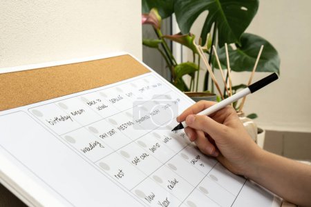 Photo for Female hand writing with marker on Monthly PLANNER filled with appointments and plans for next month. Busy month schedule. Magnetic board with the days of the month. Place to enter important matters schedule. Concept for business planning. Whiteboard - Royalty Free Image