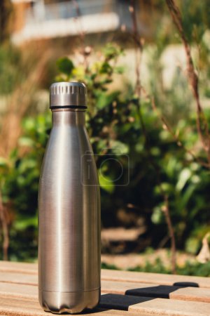Foto de Water bottle. Reusable steel thermo water bottle on wooden bench. Sustainable lifestyle. Plastic free zero waste free living. Go green Environment protection. Health-conscious. Steel thermo water bottle of silver, on background of blurred grass. - Imagen libre de derechos